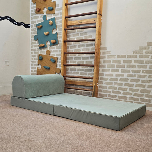 A children's play area in a sunlit room featuring a light sage green foam step stool and matching round ottoman on a beige rug, with wooden toys, a wall ladder, and puzzle decorations in the background.