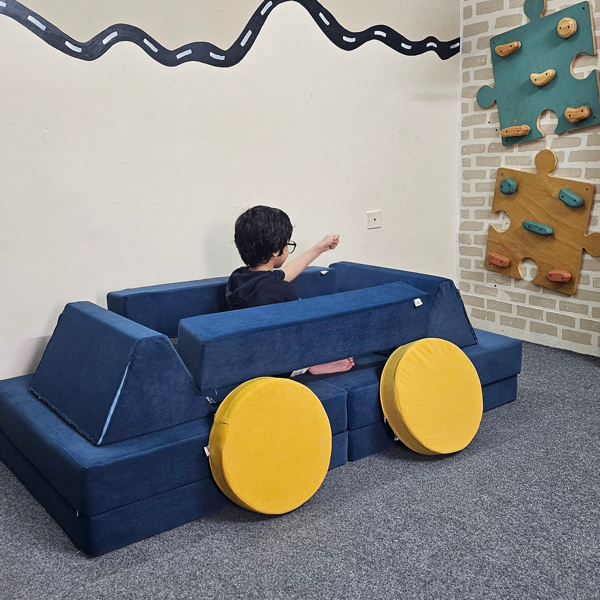 An overhead view of a child sitting on a blue padded play couch with two round yellow cushions in an indoor play area. A gray textured wall is on the right, and a white floor with a black wavy line is on the left.