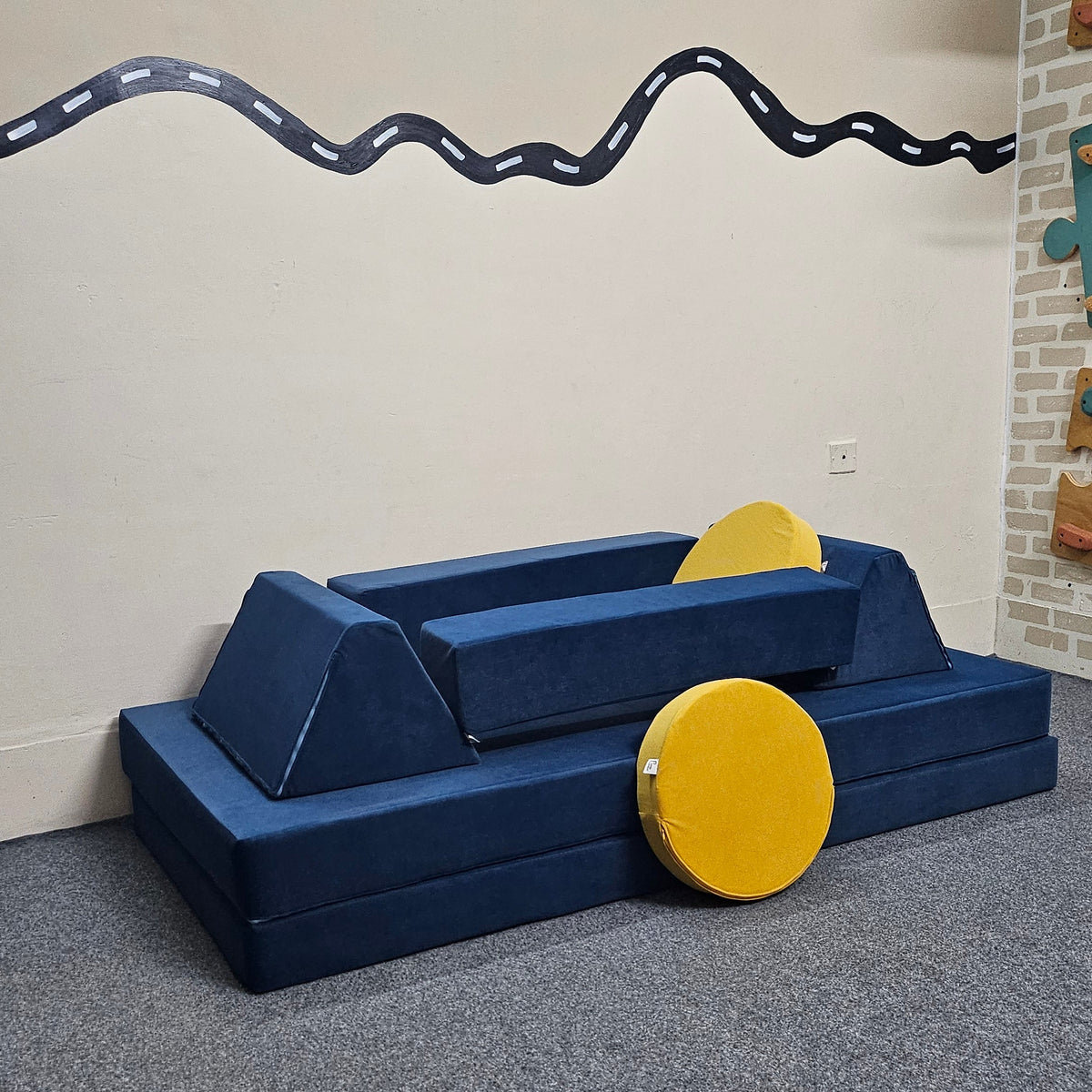 A blue foam play couch with yellow wheels and a yellow cushion, placed on a gray carpeted floor against a beige wall with a decorative wavy line.