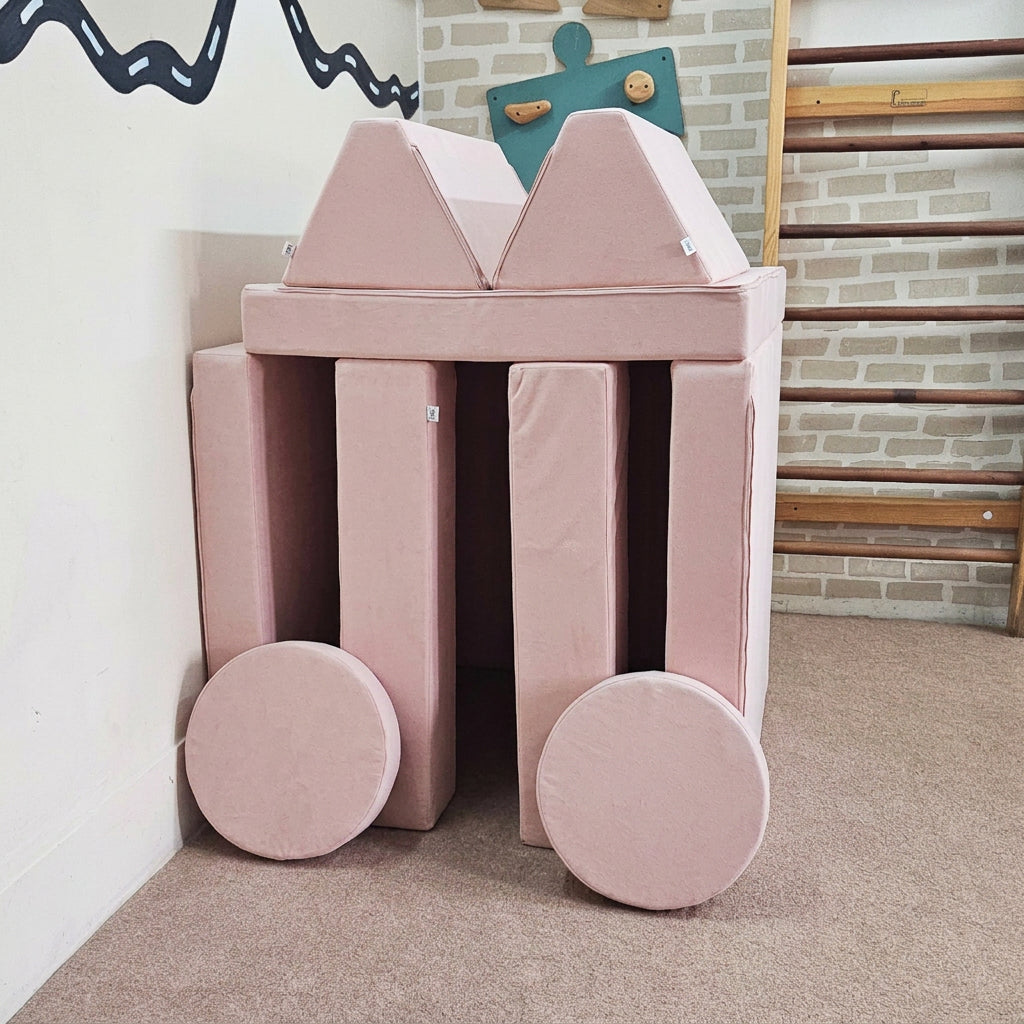 A children's foam play structure made of light blue padded blocks and wheels, set in a playroom corner with carpet, a brick-pattern wall, climbing bars, and wall art.