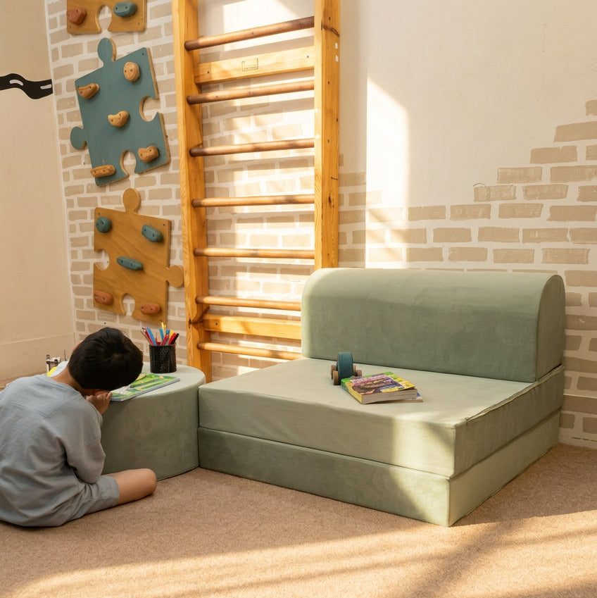A child sits on the floor beside a small modular upholstered play couch in a playroom with a wooden ladder wall, puzzle-shaped wall climbing elements, and a carpeted floor.