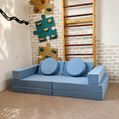 A modular light blue children's play couch with two round blue cushions, set in a room with a wooden climbing wall ladder, puzzle wall decals, and a beige carpet.