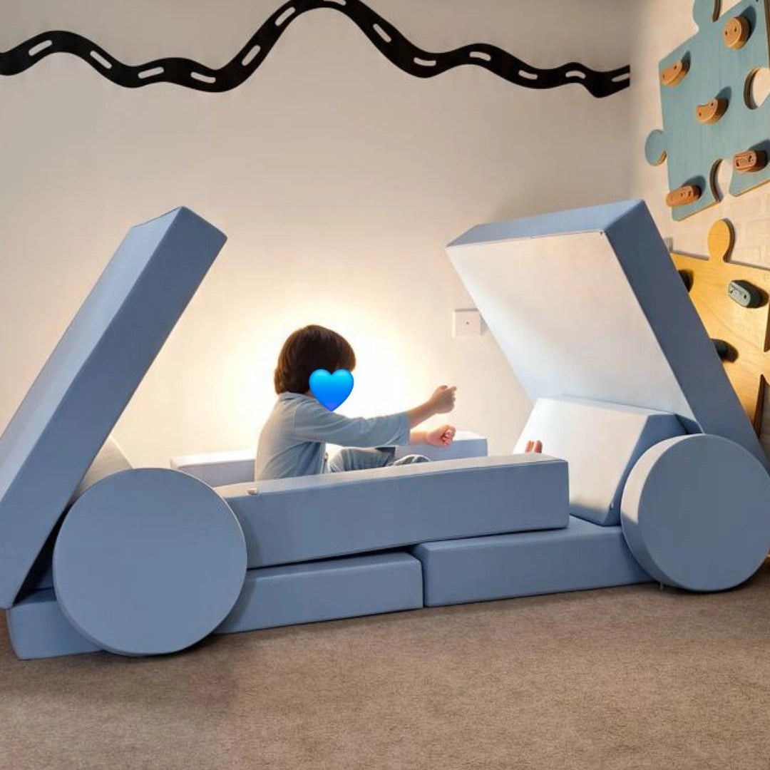 A child sits inside a large blue and white block play structure shaped like a vehicle or cart in a playroom. The room has a beige carpet, a wavy black wall decal, and a colorful puzzle wall on the right.