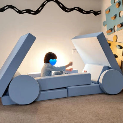 A child sits inside a large blue and white block play structure shaped like a vehicle or cart in a playroom. The room has a beige carpet, a wavy black wall decal, and a colorful puzzle wall on the right.